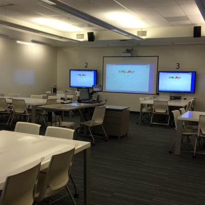 3 monitors on main wall, one monitor is visible on left wall. Table and chairs in groups.